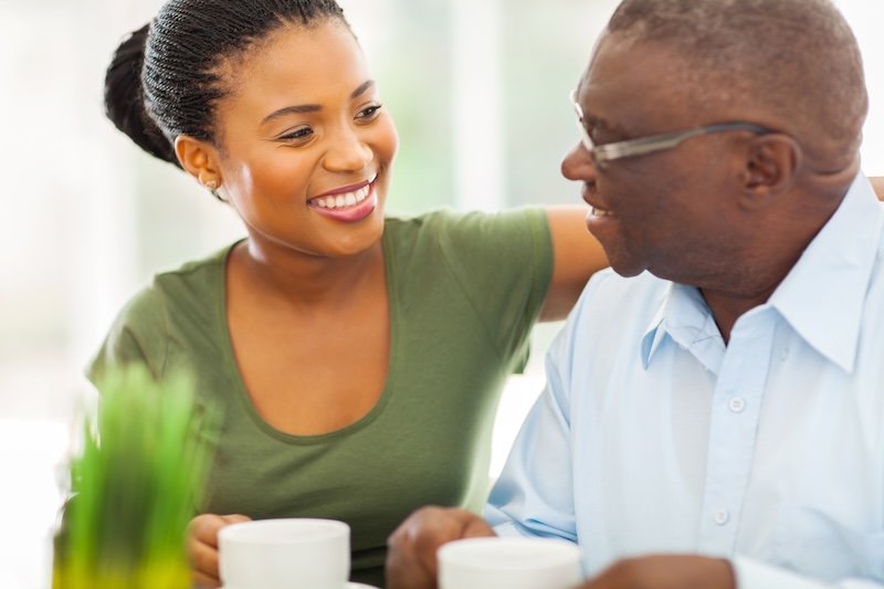 A caregiver providing 24-hour live-in care smiles as she puts her arm around an older man with dementia.